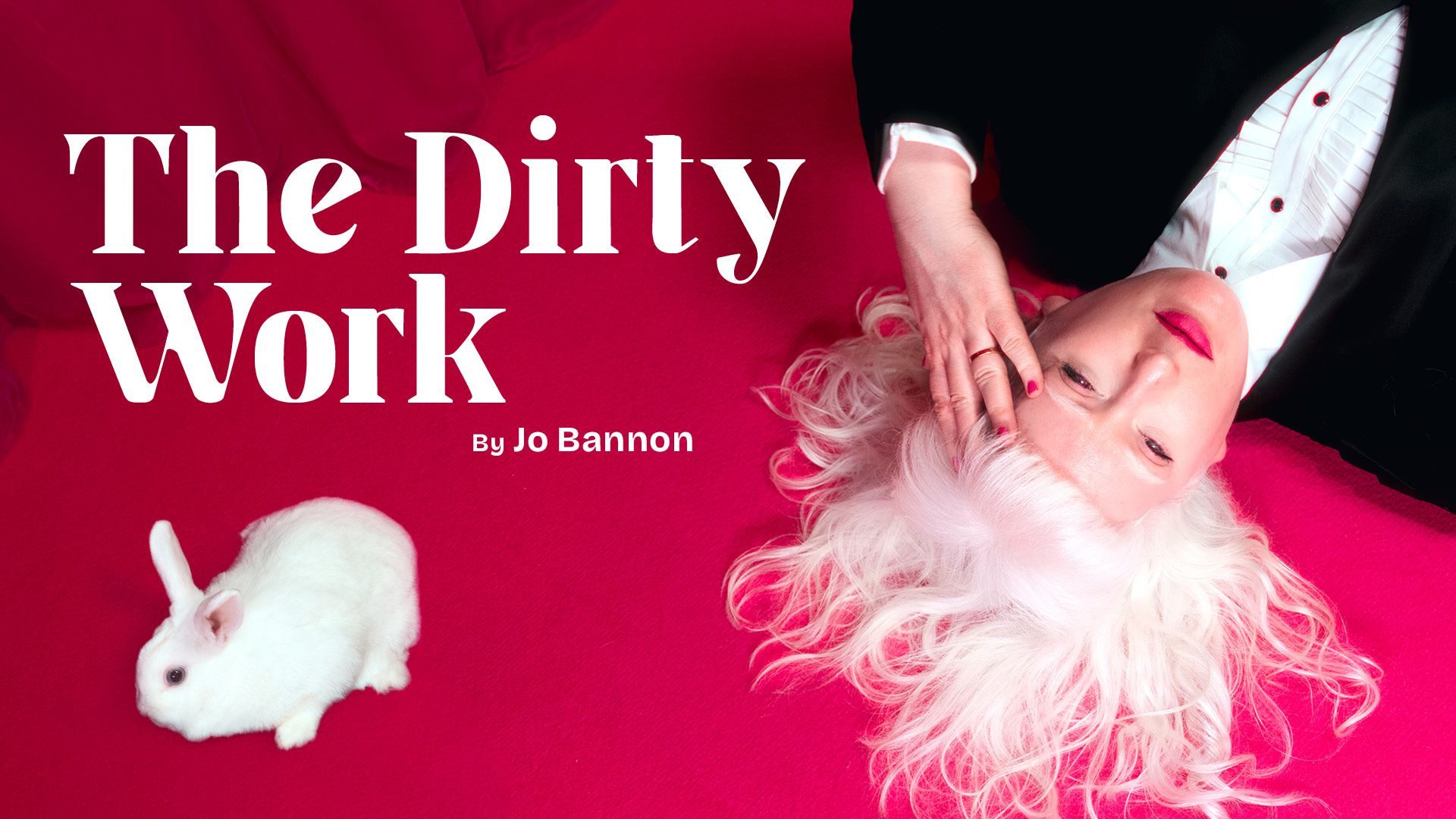 A photograph of Jo Bannon lying on a vibrant pink carpet with her white hair fanned out around her head. She is wearing a black suit and white shirt and hot pink lipstick. Her hand is held to her forehead. A small white rabbit is positioned nearby. The background features a rich pink curtain.